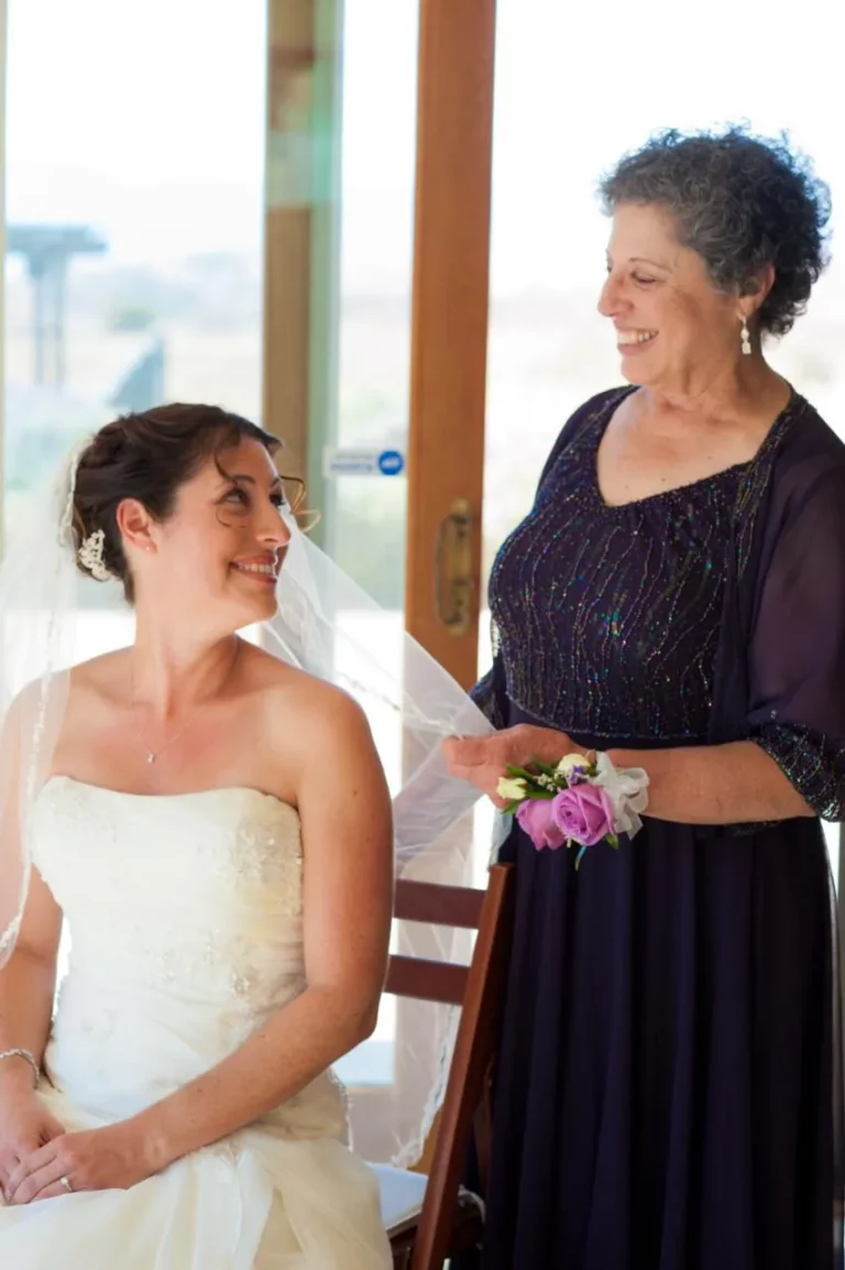 A bride and her mother share a joyful moment at a wedding, smiling at each other lovingly.